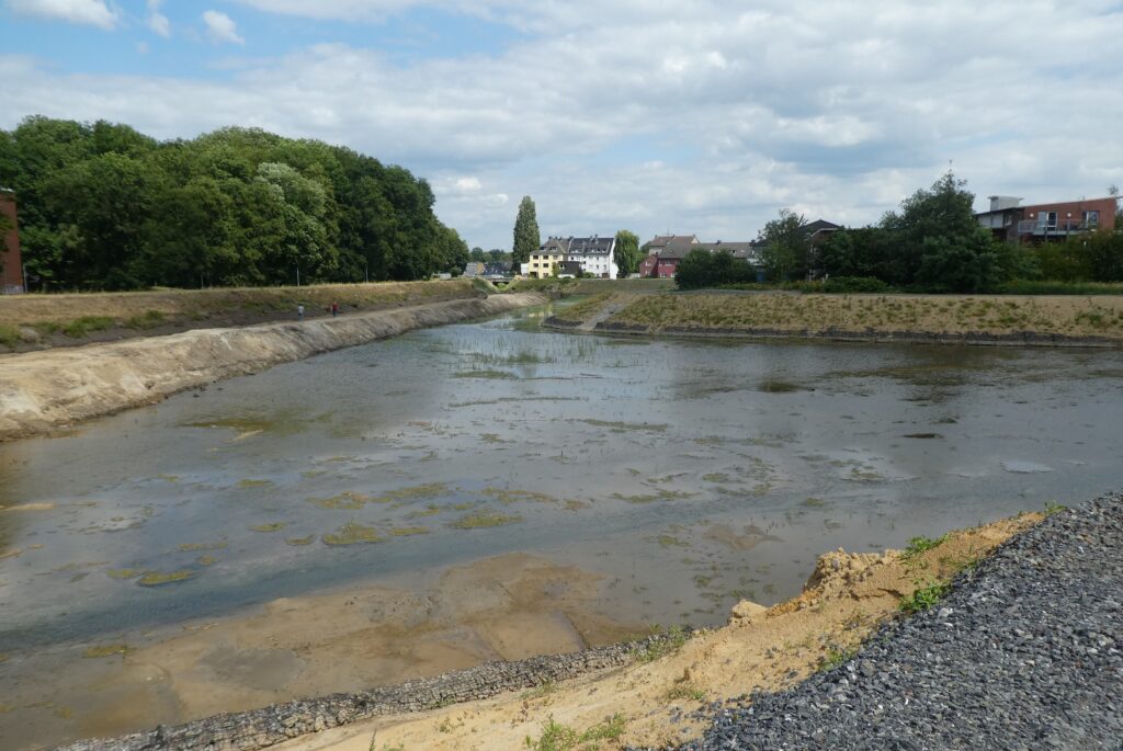 Blick entlang der Gewässeraufweitung am Dattelner Mühlenbach mit flachem Wasser, Böschungen und angrenzender Wohnbebauung.